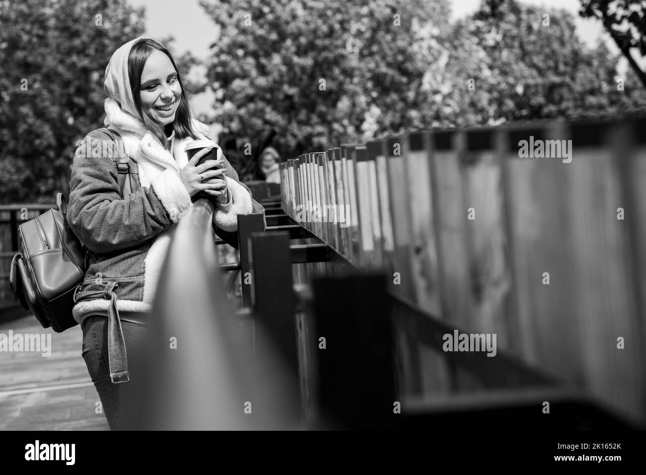 Giovane donna positiva in abiti casual con tazza di carta di caffè in piedi sulla passerella nel parco cittadino. Bianco e nero Foto Stock