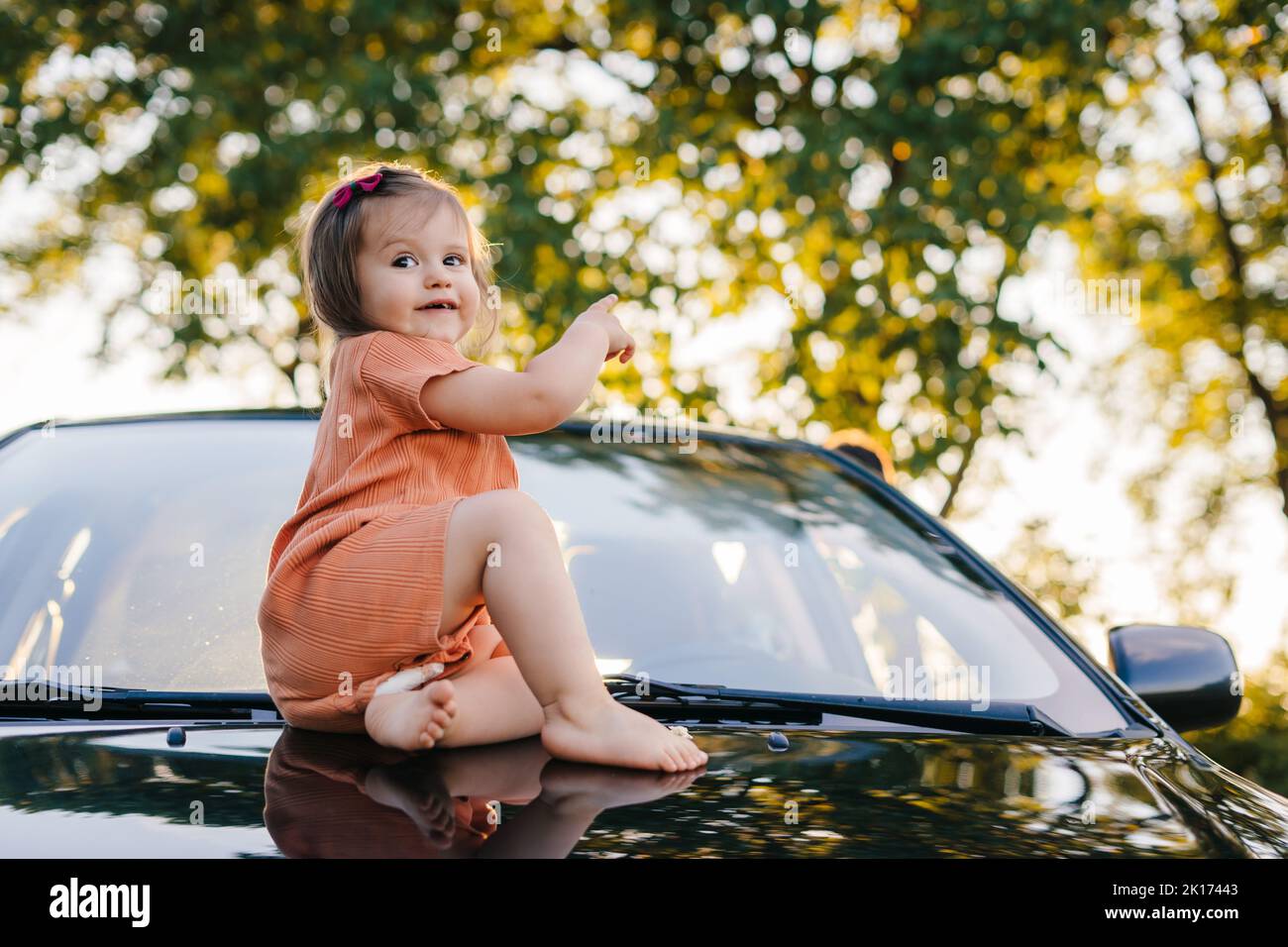 Ritratto di una bambina che si rilassa sul cappuccio dell'auto dei suoi genitori che ha fatto una sosta sulla strada durante il loro viaggio. Vacanze estive. Giorno di sole. Famiglia felice Foto Stock