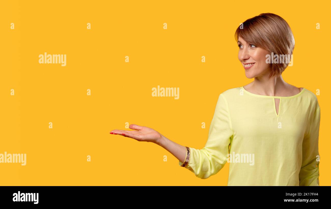 Offerta speciale. Modo di soluzione. Sfondo pubblicitario. Donna matura allegra in blusa gialla che consiglia un prodotto invisibile virtuale con palma aperta Foto Stock