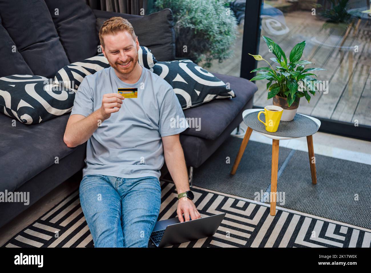 Adorabile uomo adulto bearded, che assicura le informazioni della sua carta di credito sul suo laptop, seduto sul pavimento a casa, bere caffè. Foto Stock