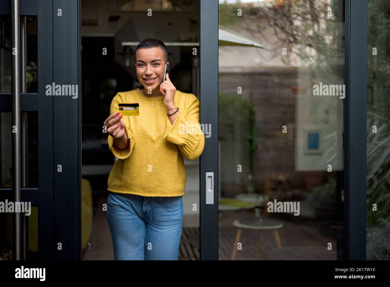 Donna adulta sorridente dai capelli corti, in piedi sulla terrazza, usando il telefono, controllando il numero della carta di credito. Foto Stock