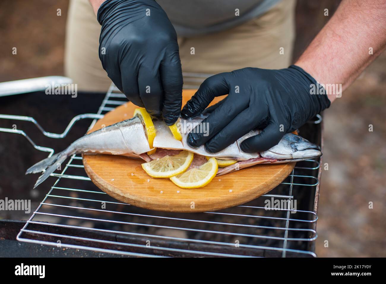 Sgombro alla griglia con fettine di limone, vista dall'alto. Il processo di cottura del pesce all'aperto. Chef in guanti in lattice nero preparazione di sgombro pesce su un bosco Foto Stock