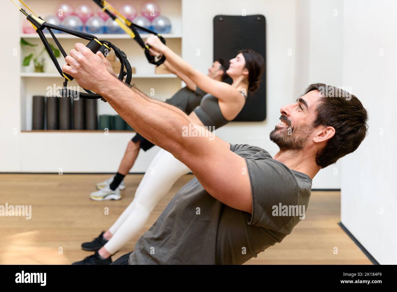 Vista laterale dell'atleta maschile bearded sorridendo e facendo l'esercizio in bassa fila con le corde TRX durante l'allenamento in sospensione di gruppo in palestra Foto Stock