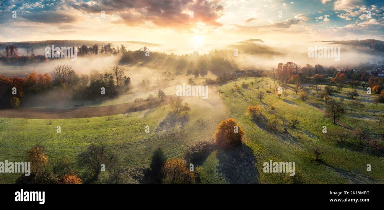 Magnifico scenario di alba aerea che mostra un paesaggio rurale, con prati, campi, colline, alberi, e bei raggi solari che cadono attraverso la nebbia Foto Stock