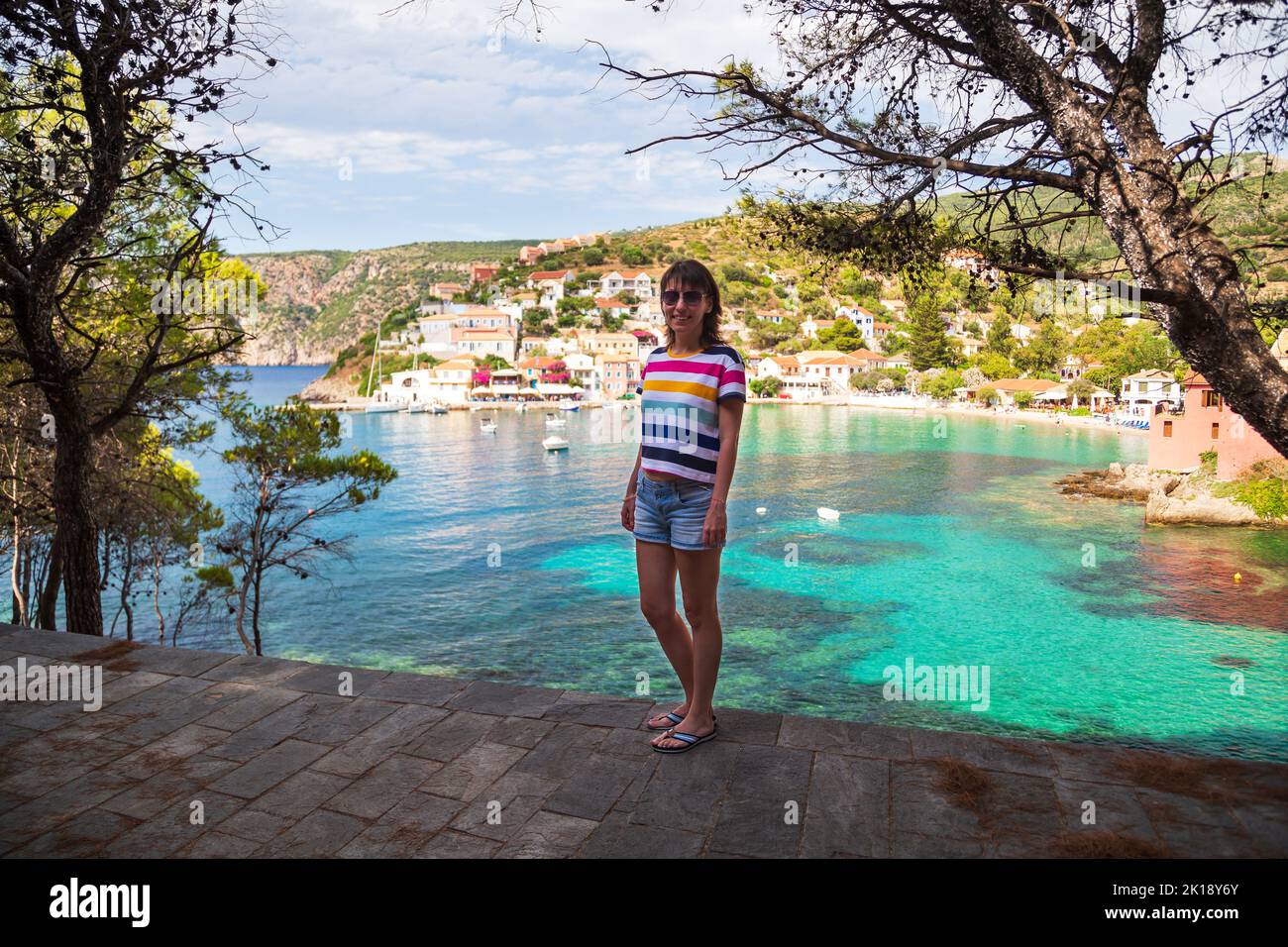 Giovane donna sorridente in camicia multicolore a righe e pantaloncini jeans nel parco verde del villaggio di Asos, nel periodo estivo. Destinazione di viaggio estremamente popolare in Foto Stock