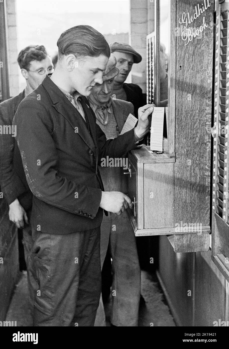 1930s, storico, operai della segheria che lavorano presso la British Coated Board & Paper Mills Ltd, South Wales, UK, utilizzando un registratore orario Gledhill Brook con una cassa in quercia. La figura mostra i lavoratori in coda, con un giovane lavoratore maschio che utilizza una chiave e una scheda sulla macchina di registrazione degli orari. Una società britannica, con sede a Huddersfield, Gledhill Brooks produsse una gamma di registratori di tempo di alta qualità fino a quando l'azienda non fu rilevata nel 1964. Foto Stock