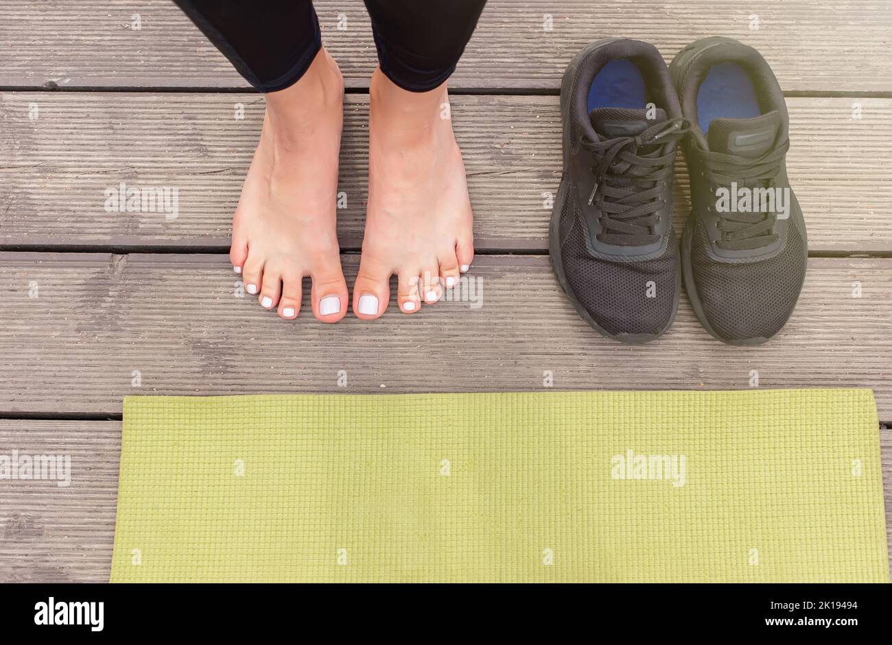 Primo piano di piedi nudi su un pavimento di legno vicino a un tappeto sportivo verde, ci sono sneakers neri nelle vicinanze Foto Stock
