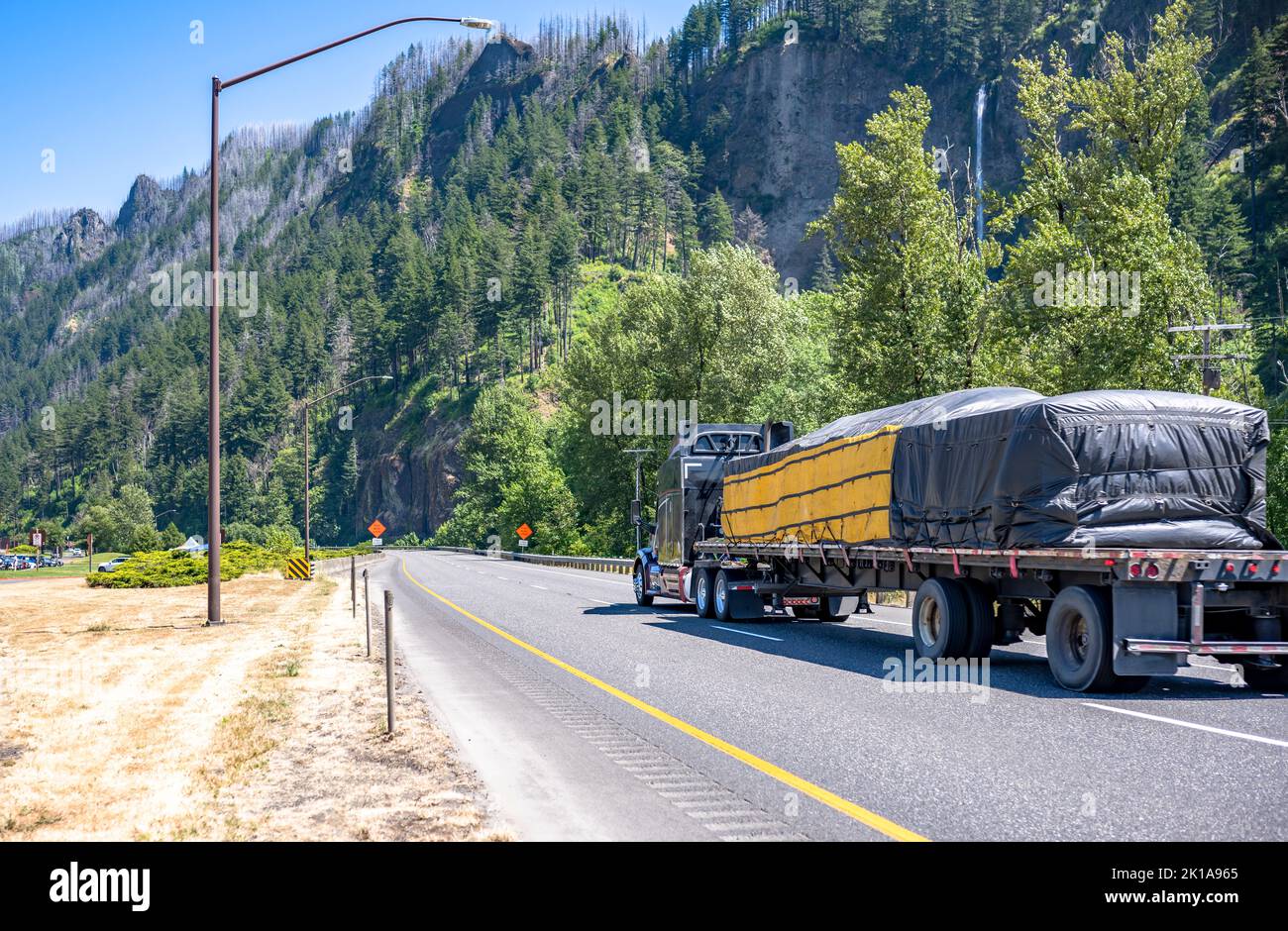 Industriale grande carro di perforazione blu classico semirimorchio che trasporta il carico coperto fissato sul semirimorchio piano che guida su strada con la montagna Foto Stock