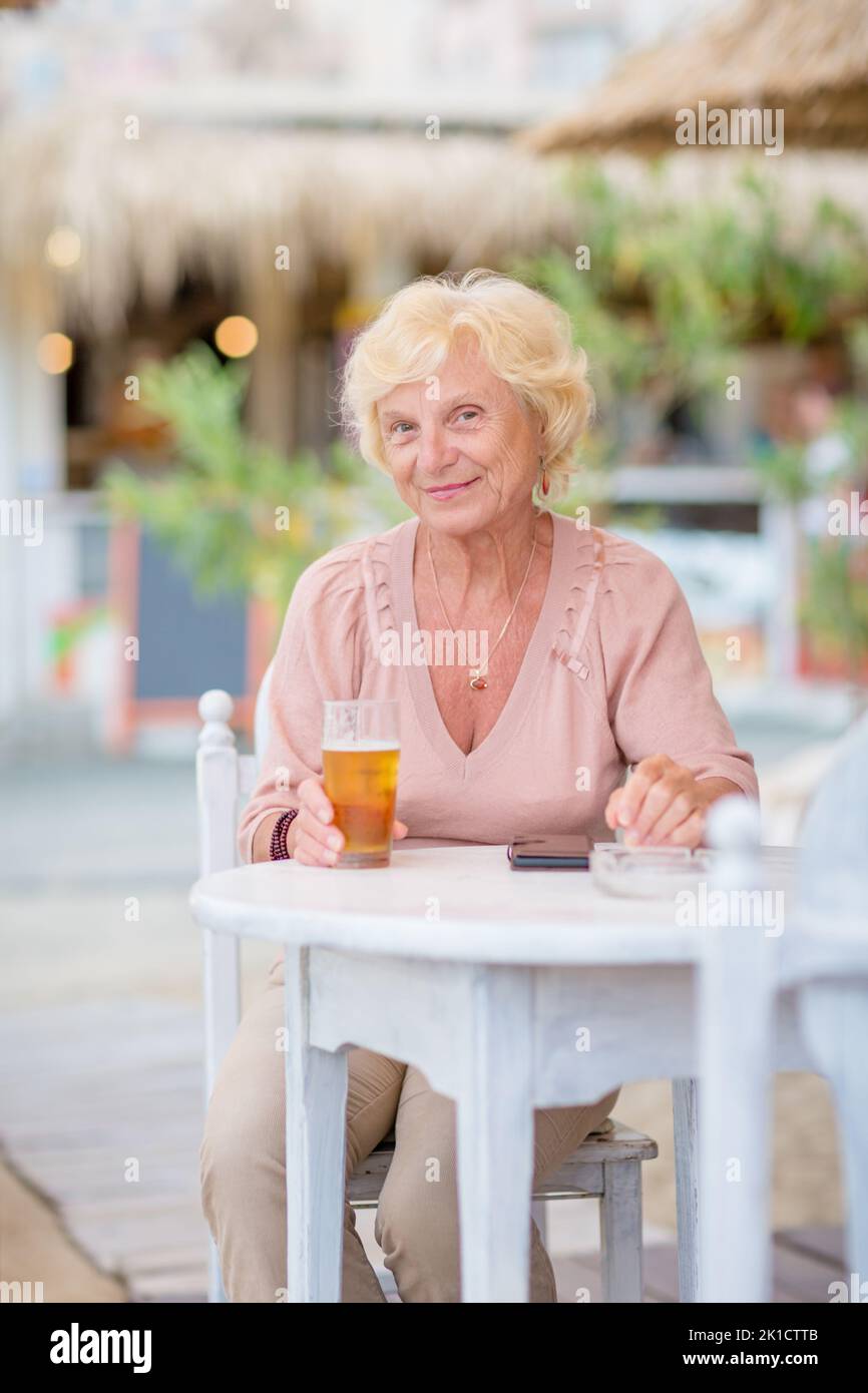 Donna matura seduta a un tavolo in un bar estivo e bere birra Foto Stock