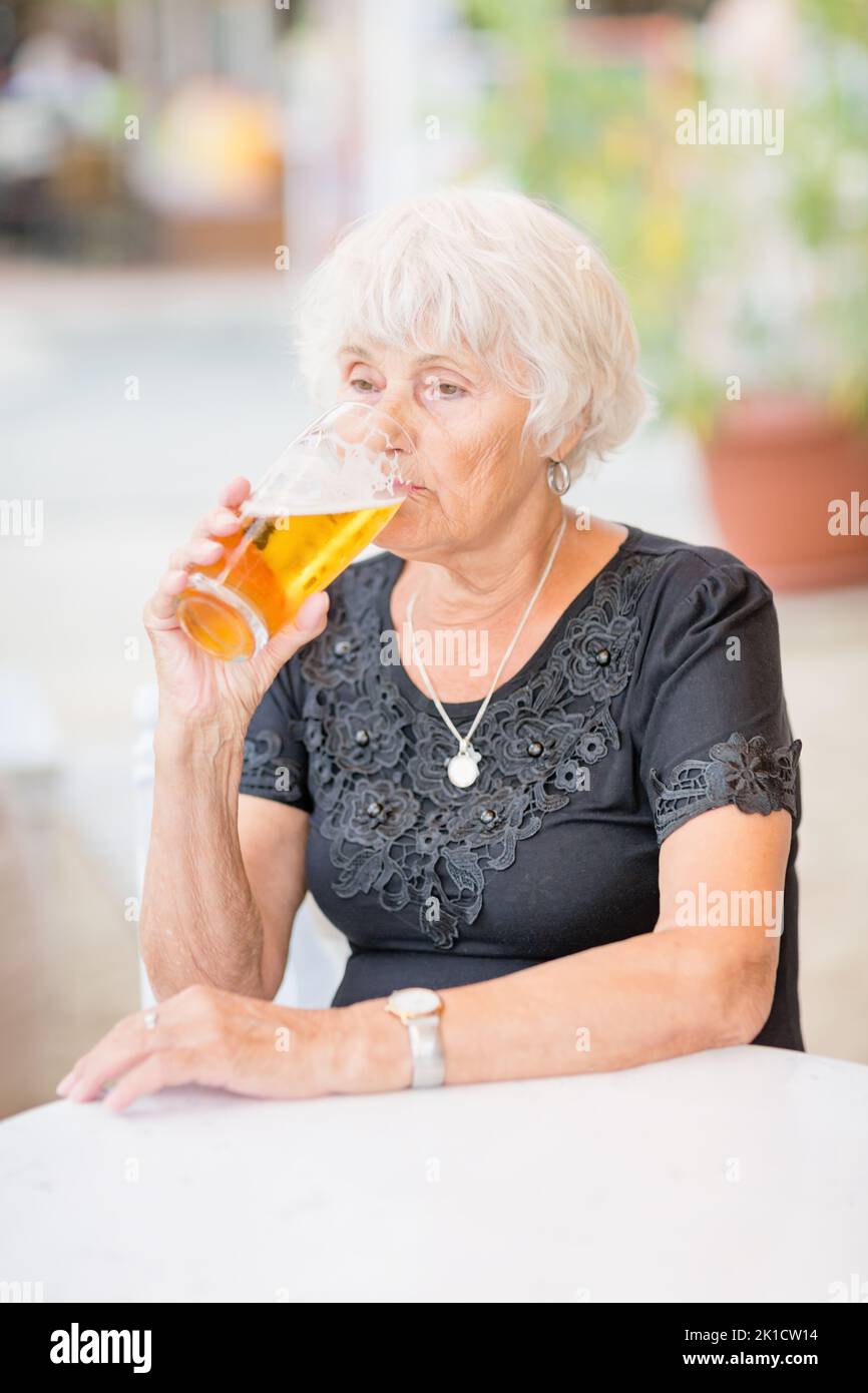 Donna matura seduta a un tavolo in un bar estivo e bere birra Foto Stock