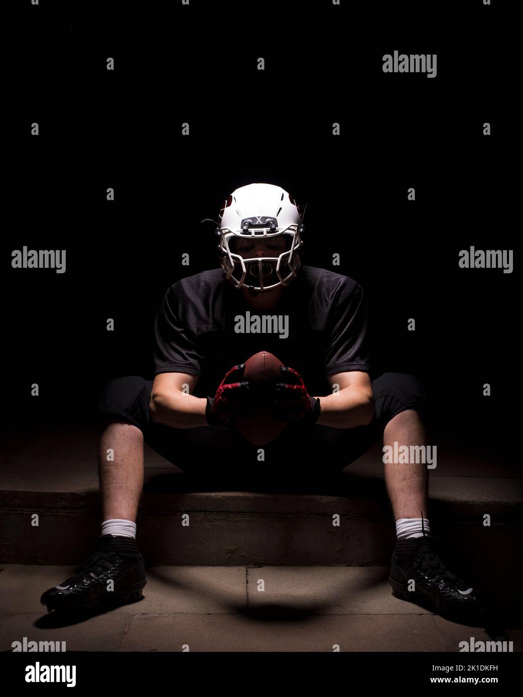 Uomo in abbigliamento da calcio americano seduto su un gradino e tenendo la palla con entrambe le mani. Foto Stock