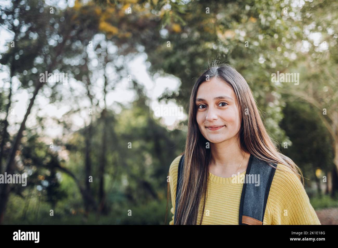 Sorridente studentessa universitaria che indossa un maglione giallo e uno zaino nella strada del parco del campus. Sorriso innocente Foto Stock