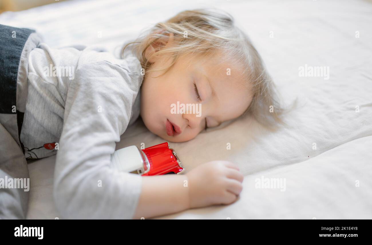 Adorabile bambino che dorme a letto con la sua macchina giocattolo. Bambino stanco che prende un pisolino sotto coperta bianca. Andare a letto per i bambini. Foto Stock