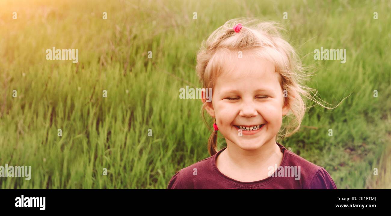 La bambina fa volti divertenti e chiude gli occhi. Spazio di copia. Allegro bambino all'aperto. Verde sfondo erba. Foto Stock