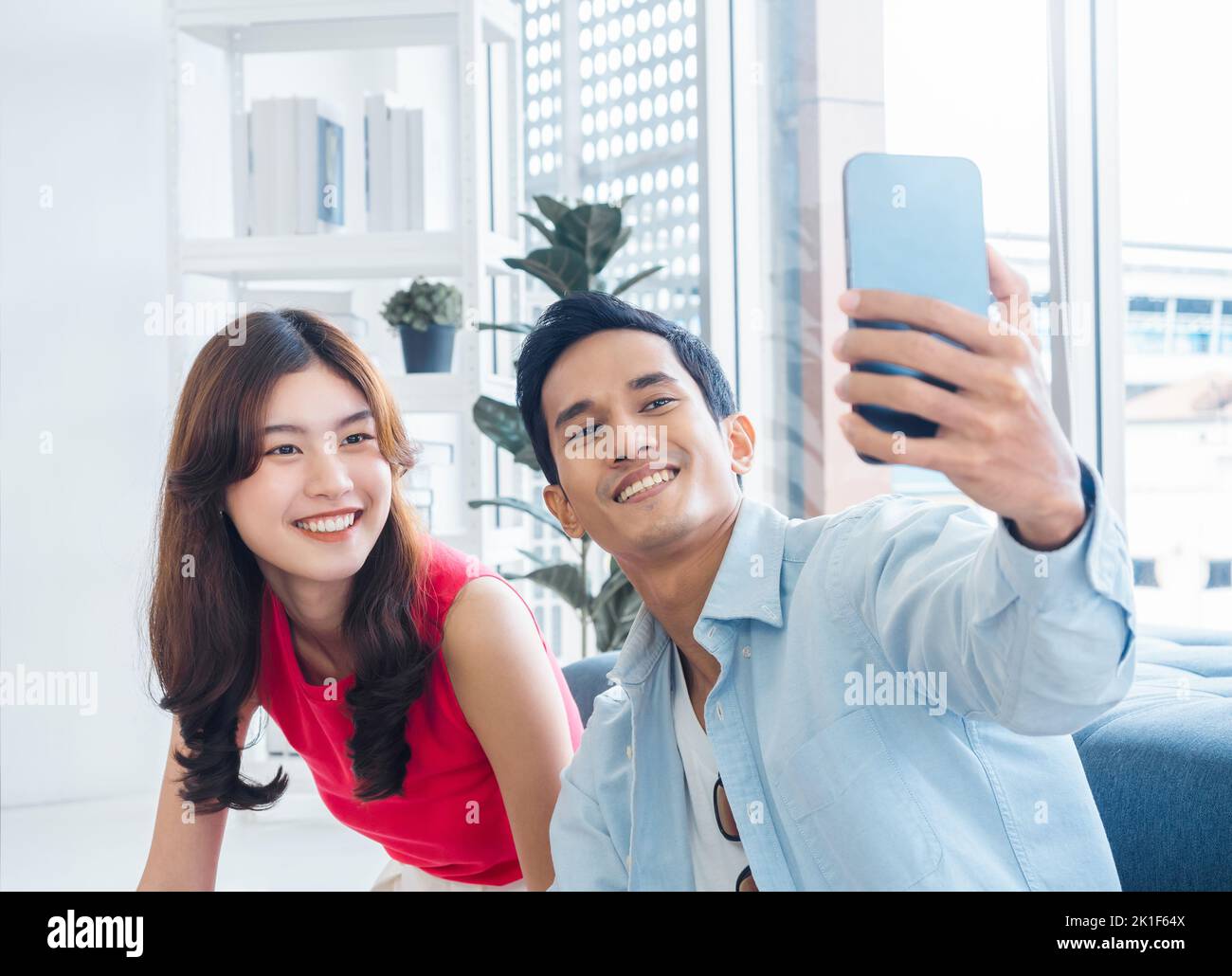 Felice bell'uomo asiatico e giovane bella donna selfie mentre si siede vicino alla finestra di vetro nel soggiorno bianco in alto edificio. Coppia allegra t Foto Stock