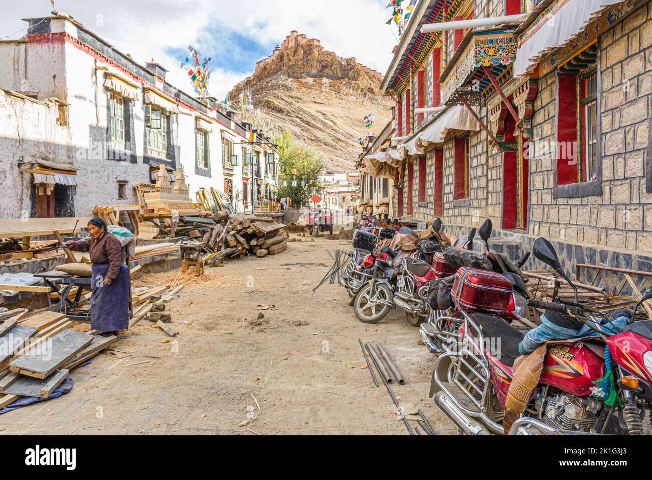 Una strada laterale nella città di Gyantse, nella prefettura di Shigatse, Cina del Tibet con una fortezza di Dzong sullo sfondo e una donna che lavora sulla strada Foto Stock