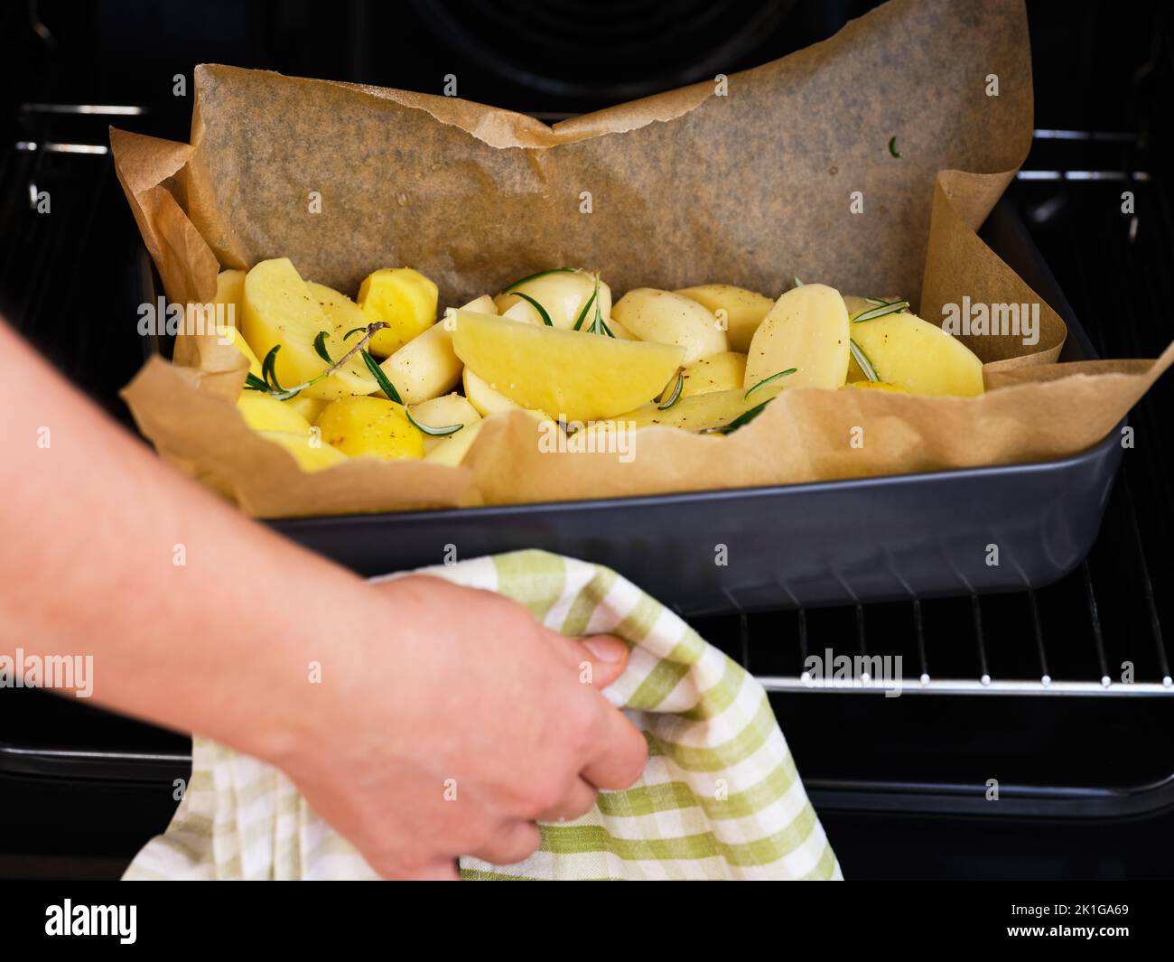 Una donna che mette in un forno una teglia nera con patate crude con rosmarino e spezie. Foto Stock