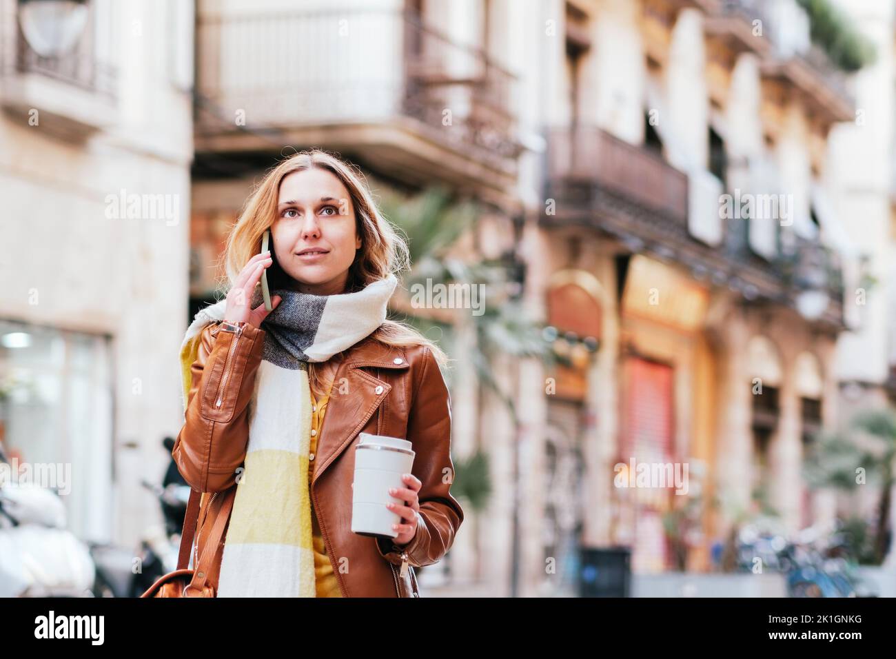 Affascinante donna con caffè da asporto che guarda e parla sul cellulare mentre si trova in piedi sulla strada con edifici residenziali in città Foto Stock