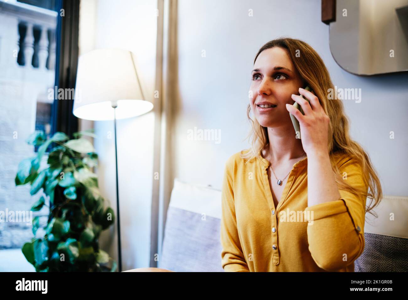 giovane donna bionda con camicia gialla seduta in una caffetteria e che parla per telefono Foto Stock