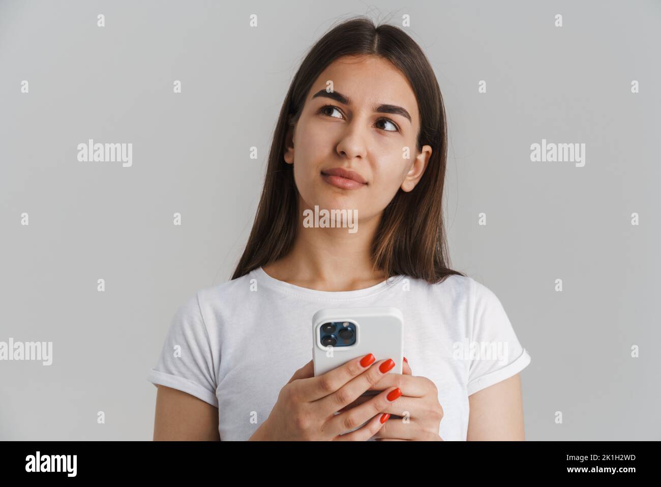 Ritratto di una giovane donna bianca casual pensiva in t-shirt con lunghi capelli brunette in piedi su sfondo grigio parete tenendo il telefono cellulare guardare asid Foto Stock