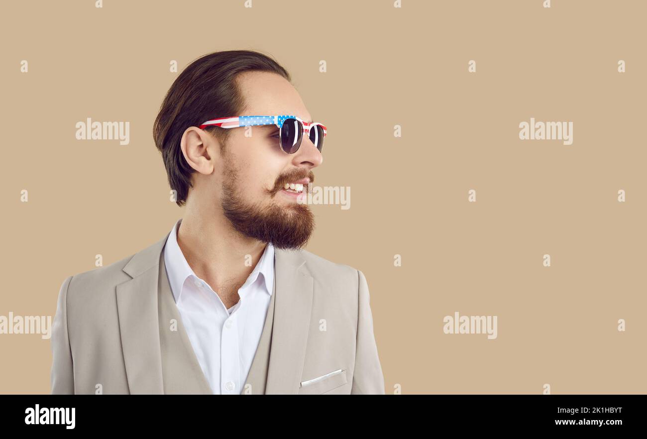 Bell'uomo fiducioso in tuta e occhiali da sole con stampa American flag sorridente su sfondo beige. Foto Stock