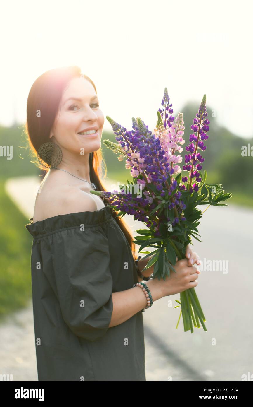 Donna attraente sulla strada in un campo estivo tra i fiori Lupinus. Foto verticale. Foto Stock