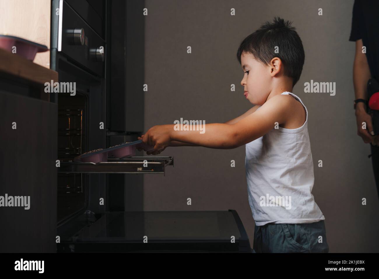 Ragazzo che cerca di mettere il vassoio di biscotti nel forno senza l'aiuto di sua madre. Stile di vita delle persone. Genitore, figlio. Famiglia felice, infanzia. Foto Stock