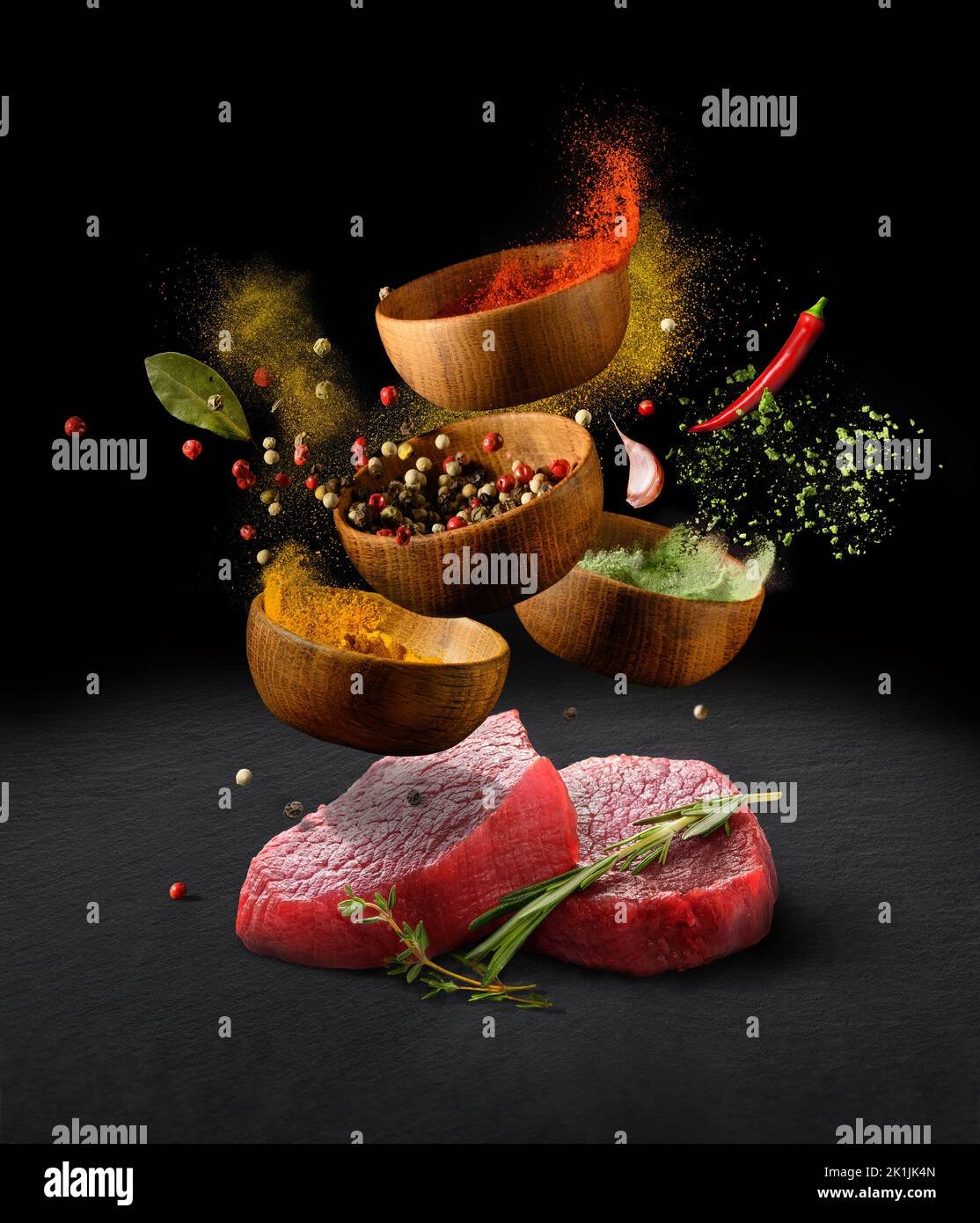Bistecche di manzo crudo su un tavolo da taglio in pietra nera e varie spezie ed erbe che volano con spruzzi su sfondo nero Foto Stock