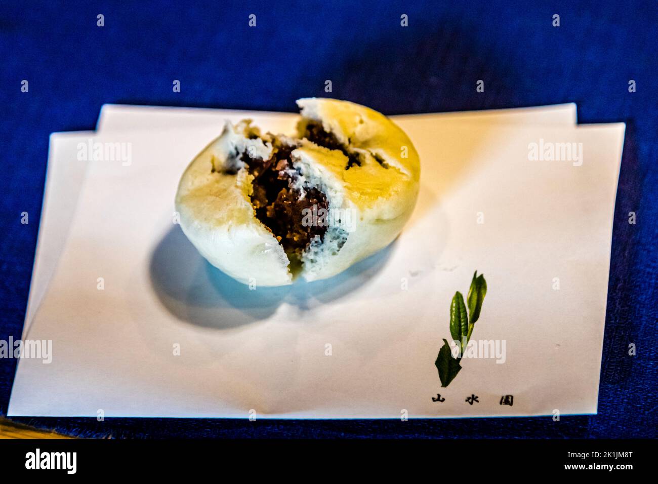 Un dolce wagashi morso, ripieno di pasta di fagioli azuki, poggia su un tovagliolo di carta stampato con il logo della fattoria del tè Sansuien vicino a Shizuoka, in Giappone – il dolce tradizionalmente apre una cerimonia del tè. Foto Stock