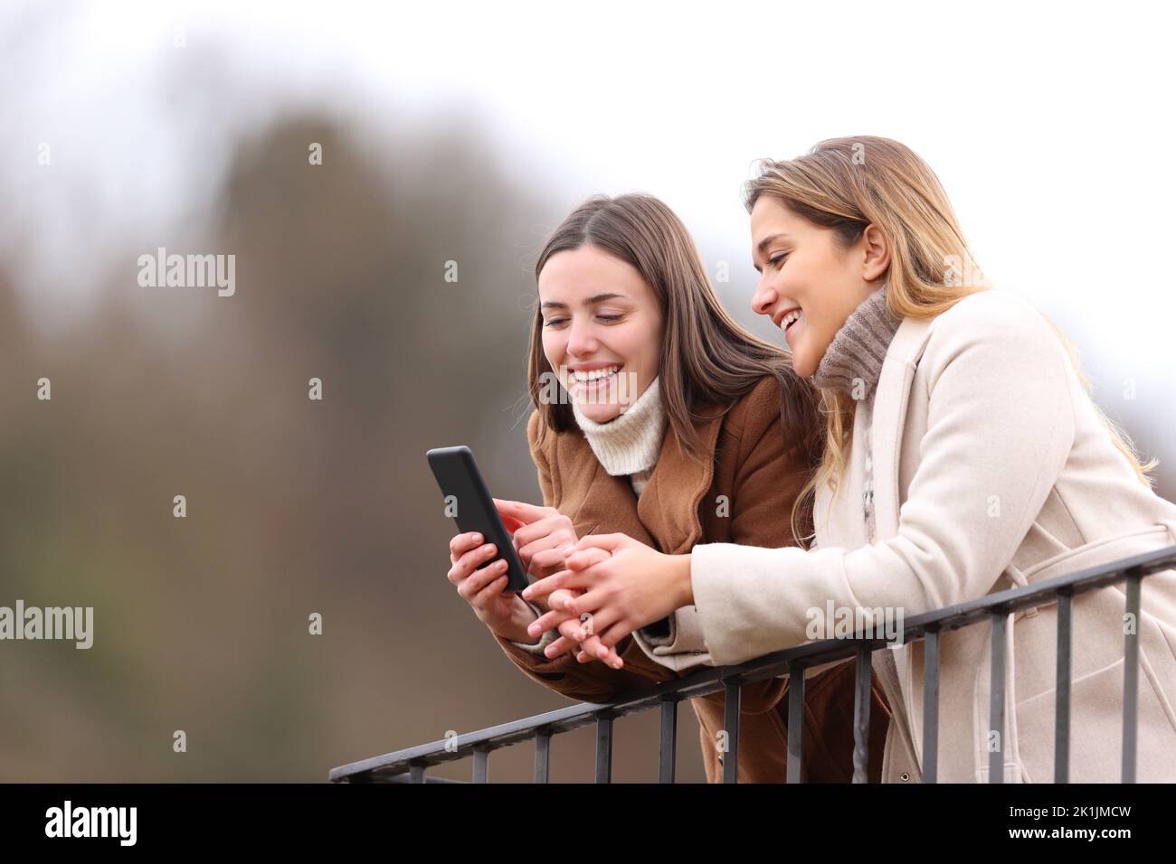 Due amici felici che controllano lo smartphone in inverno in un balcone Foto Stock