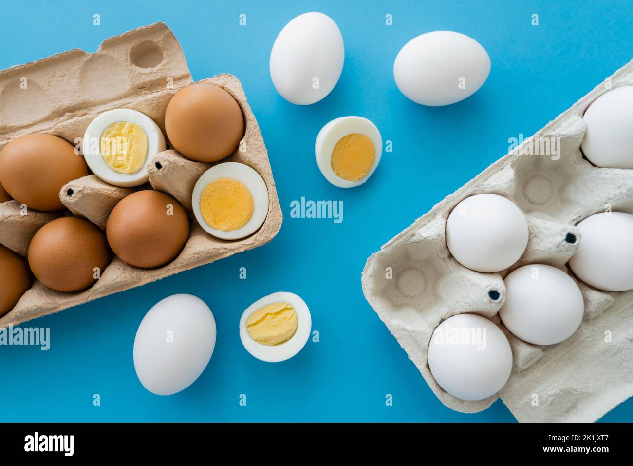 Vista dall'alto delle uova naturali crude e bollite vicino ai vassoi di cartone su sfondo blu Foto Stock