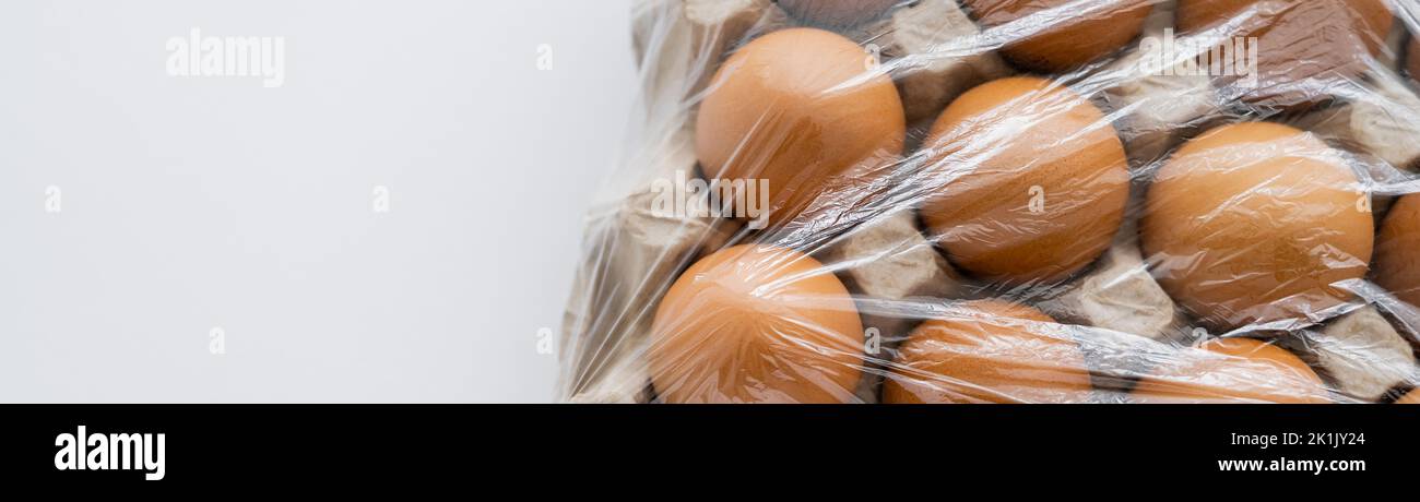 Vista dall'alto delle uova marroni sotto il cellofano in scatola di cartone su sfondo bianco, banner Foto Stock