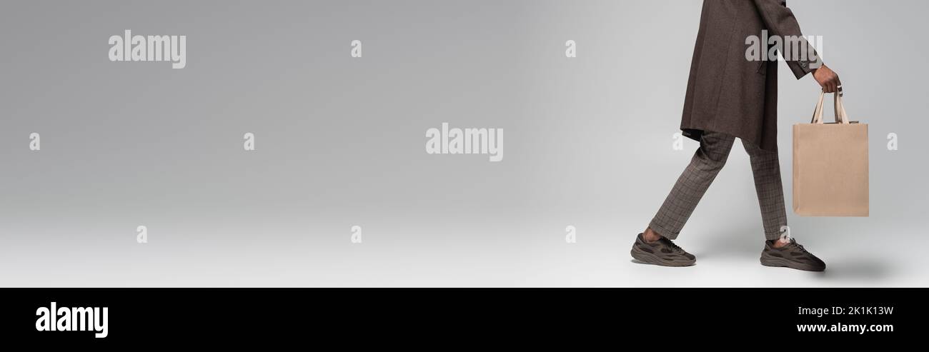 vista ritagliata dell'elegante uomo afroamericano in cappotto e sneakers camminando con borse della spesa in grigio, banner, immagine stock Foto Stock