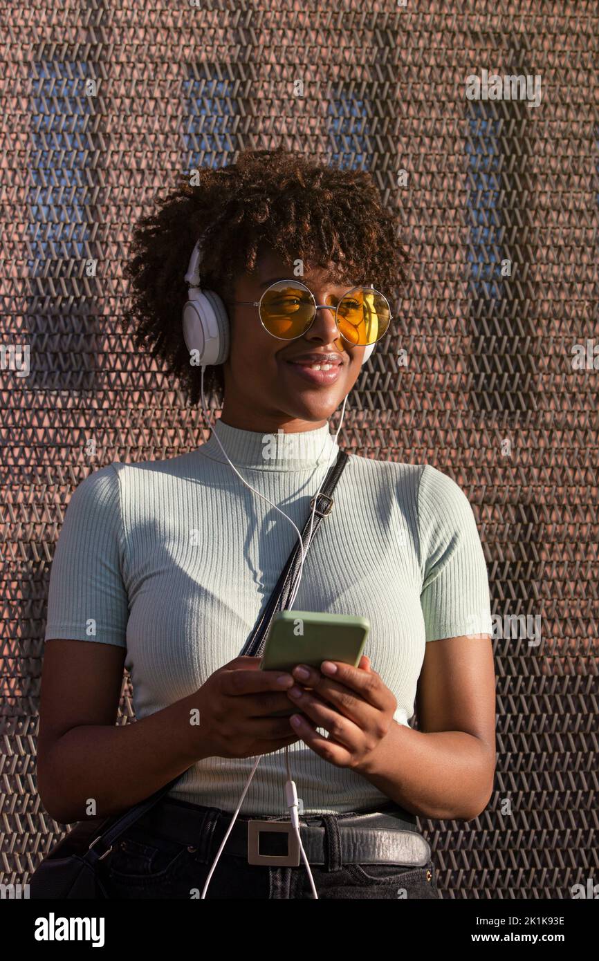 Giovane donna dell'america latina con capelli afro e occhiali sorridenti ascoltando musica su smartphone con cuffie Foto Stock