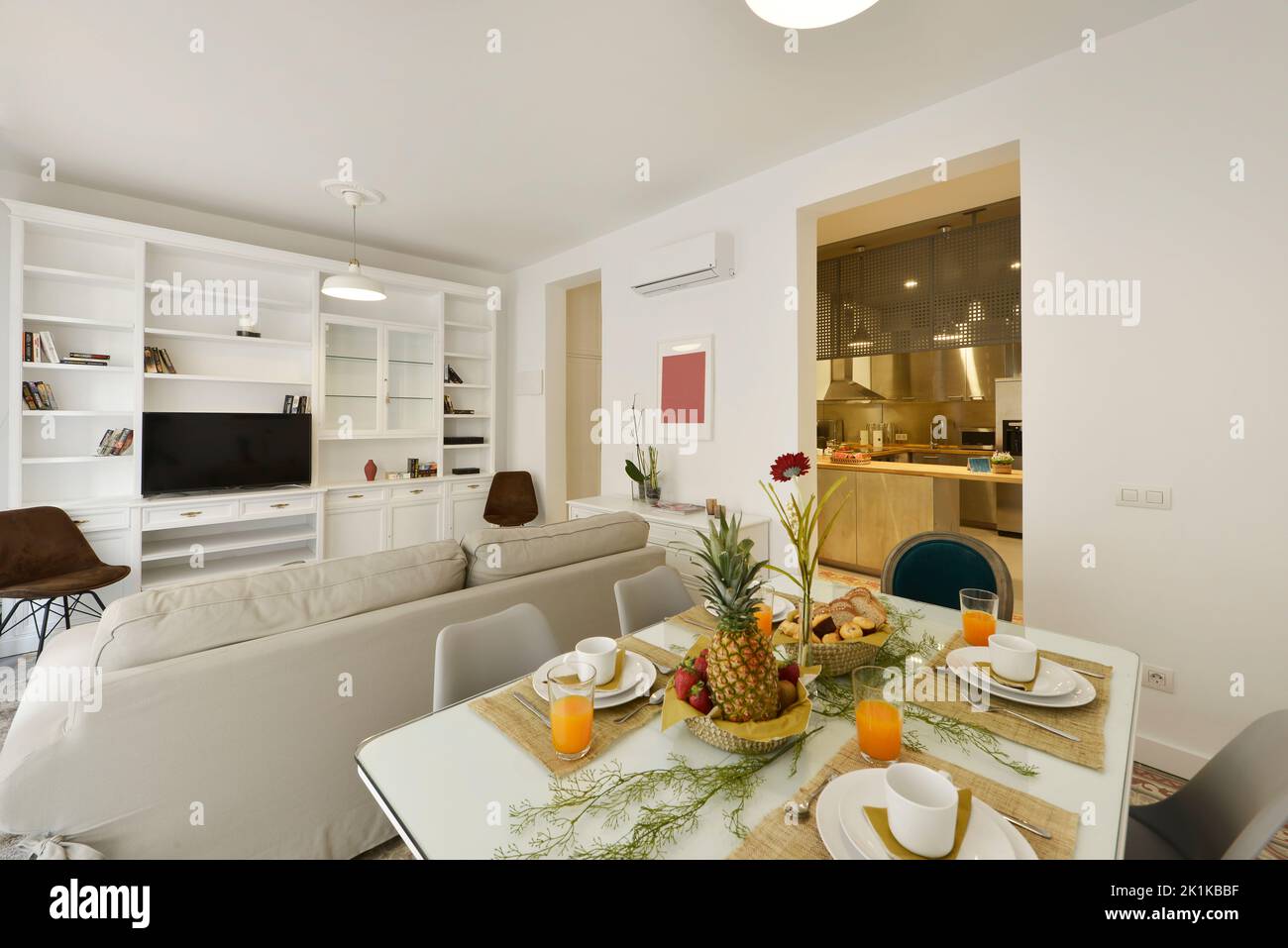 appartamento dotato di soggiorno con tavolo da pranzo in legno bianco, servizio di prima colazione con una bacinella di frutta e un vassoio di dolci e succo d''arancia in vetro Foto Stock
