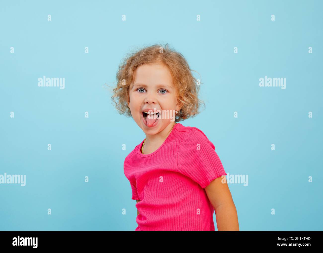 Ritratto di felice bambina con occhi blu con capelli ricci che guardano, mostrando la lingua, grimacing teasing su sfondo blu. Foto Stock