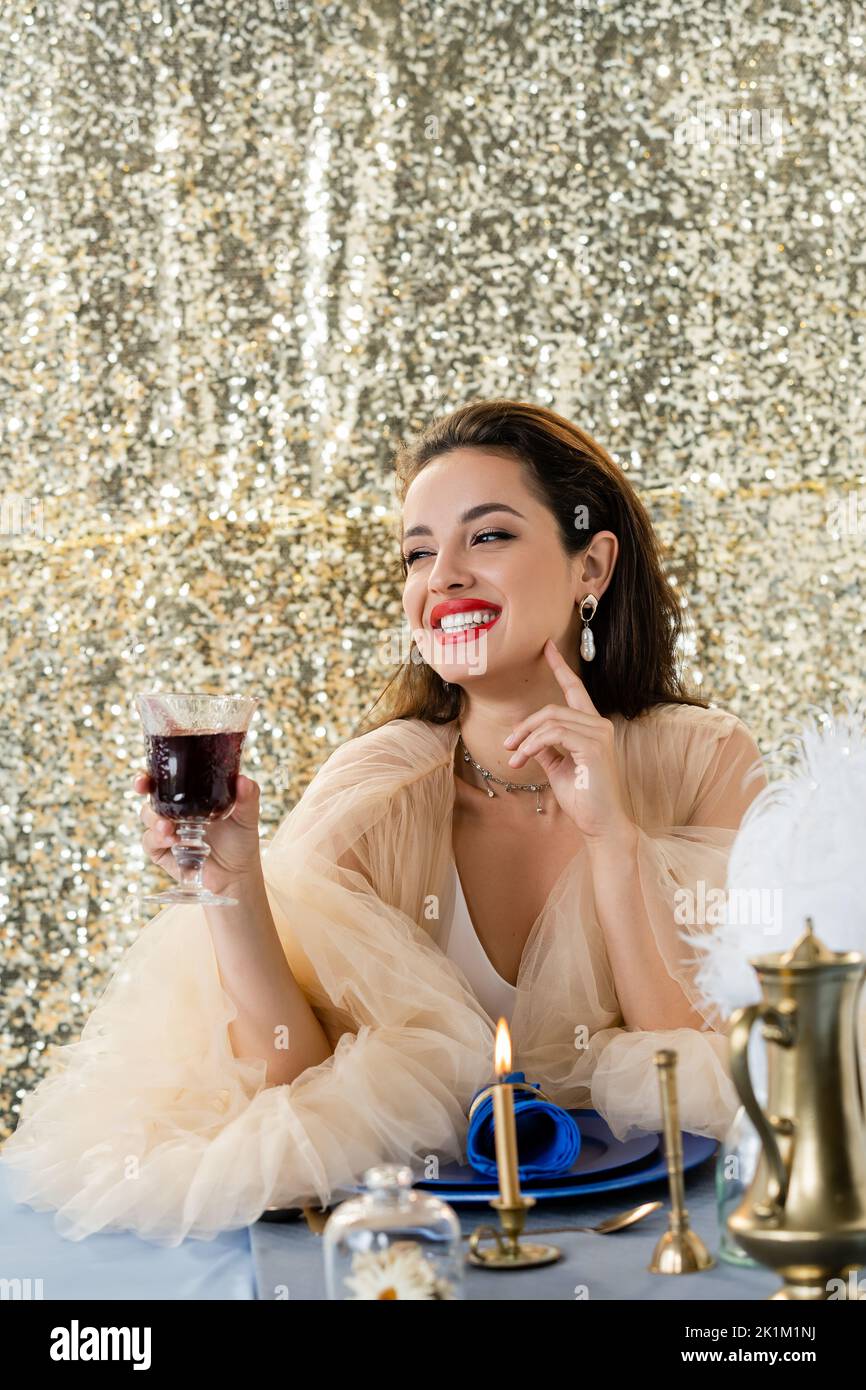 donna felice in elegante tulle vestito che tiene un bicchiere di vino rosso e guardando via su sfondo lucido, immagine stock Foto Stock