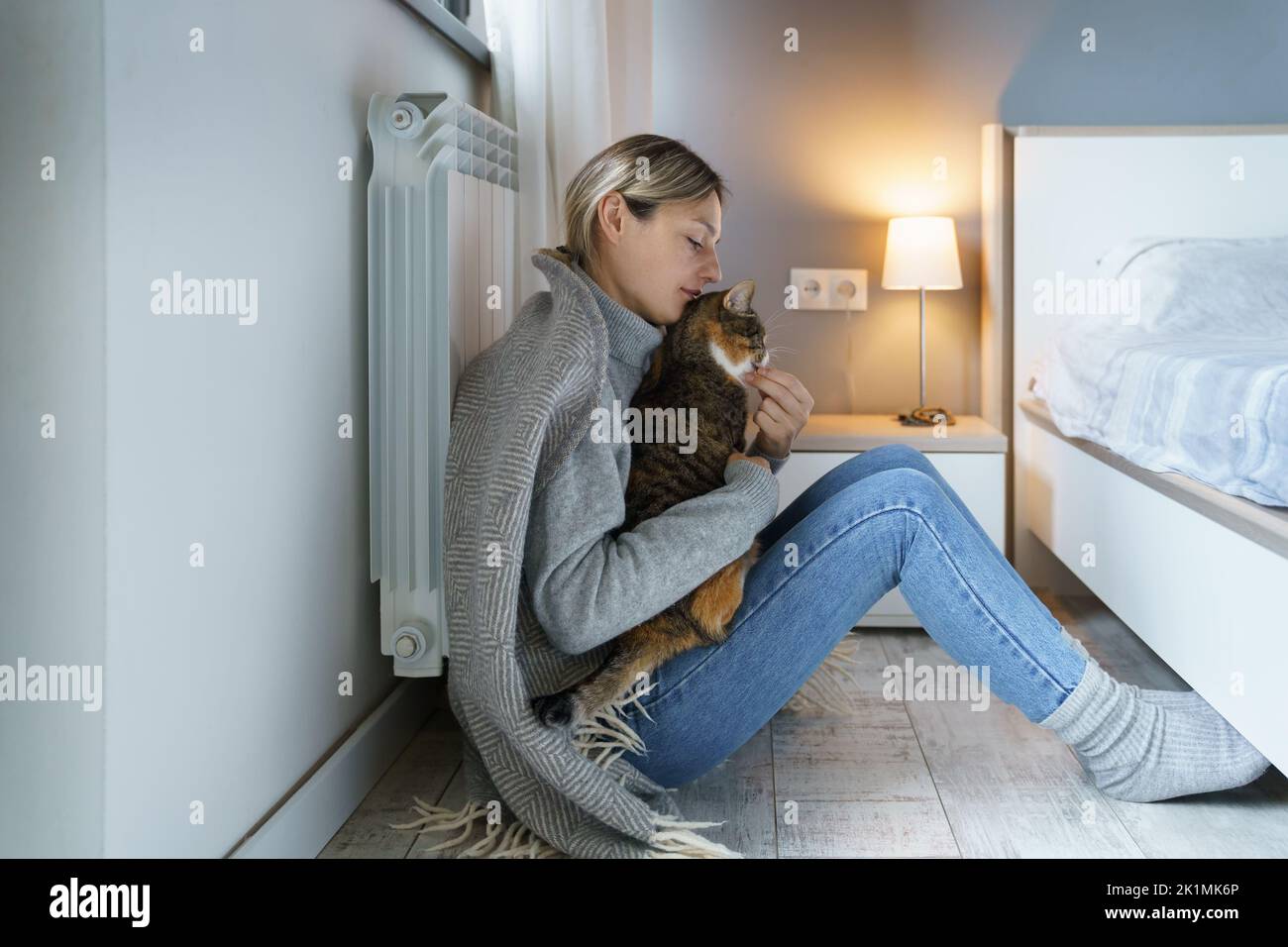 Una giovane donna coperta di coperta calda gioca con gatto che mangia animali con cibo seduto sul pavimento Foto Stock