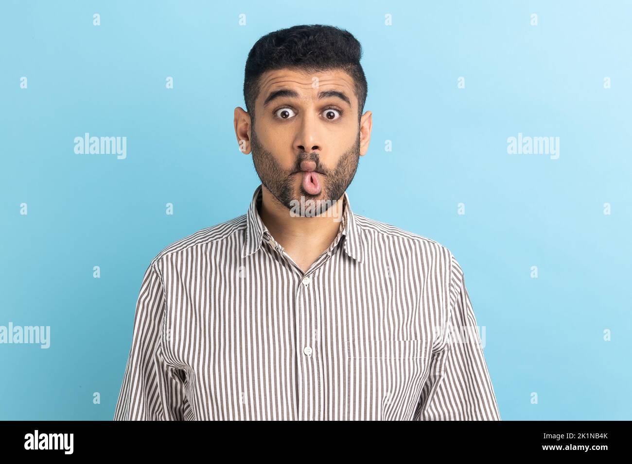 Ritratto di divertente uomo d'affari portatore di bambino facendo faccia di pesce o inviando bacio, divertendosi, scherzando, fingendo di essere stupido, indossando camicia a strisce. Studio in interni isolato su sfondo blu. Foto Stock