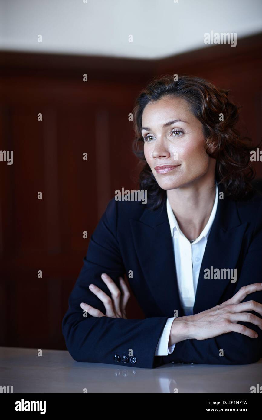 Considerando gli eventi del giorno. Donna matura avvocato guardando lontano dalla telecamera mentre si siede con le braccia ripiegate e sorridendo leggermente. Foto Stock