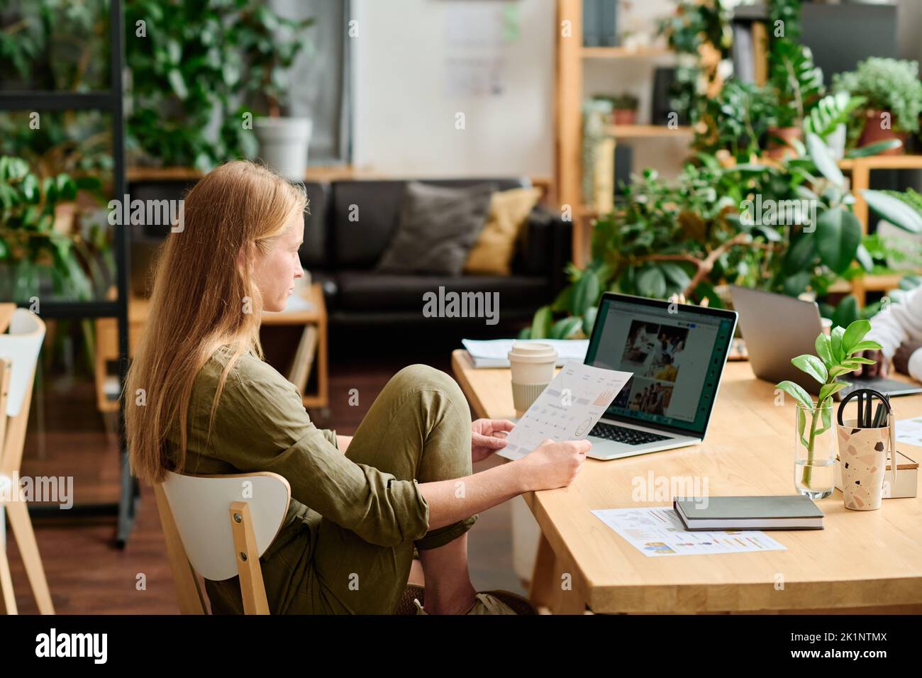 Giovane donna d'affari creativa o designer con documenti che guardano lo schermo del notebook con immagini mentre si siede sul posto di lavoro in ufficio Foto Stock
