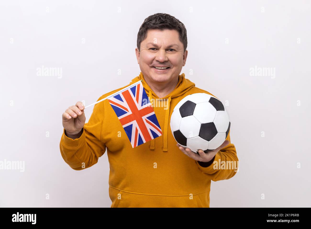 Ritratto dell'uomo soddisfatto che tiene bandiera inglese e calcio palla bianca e nera, United Football League, con felpa con cappuccio in stile urbano. Studio in interni isolato su sfondo bianco. Foto Stock