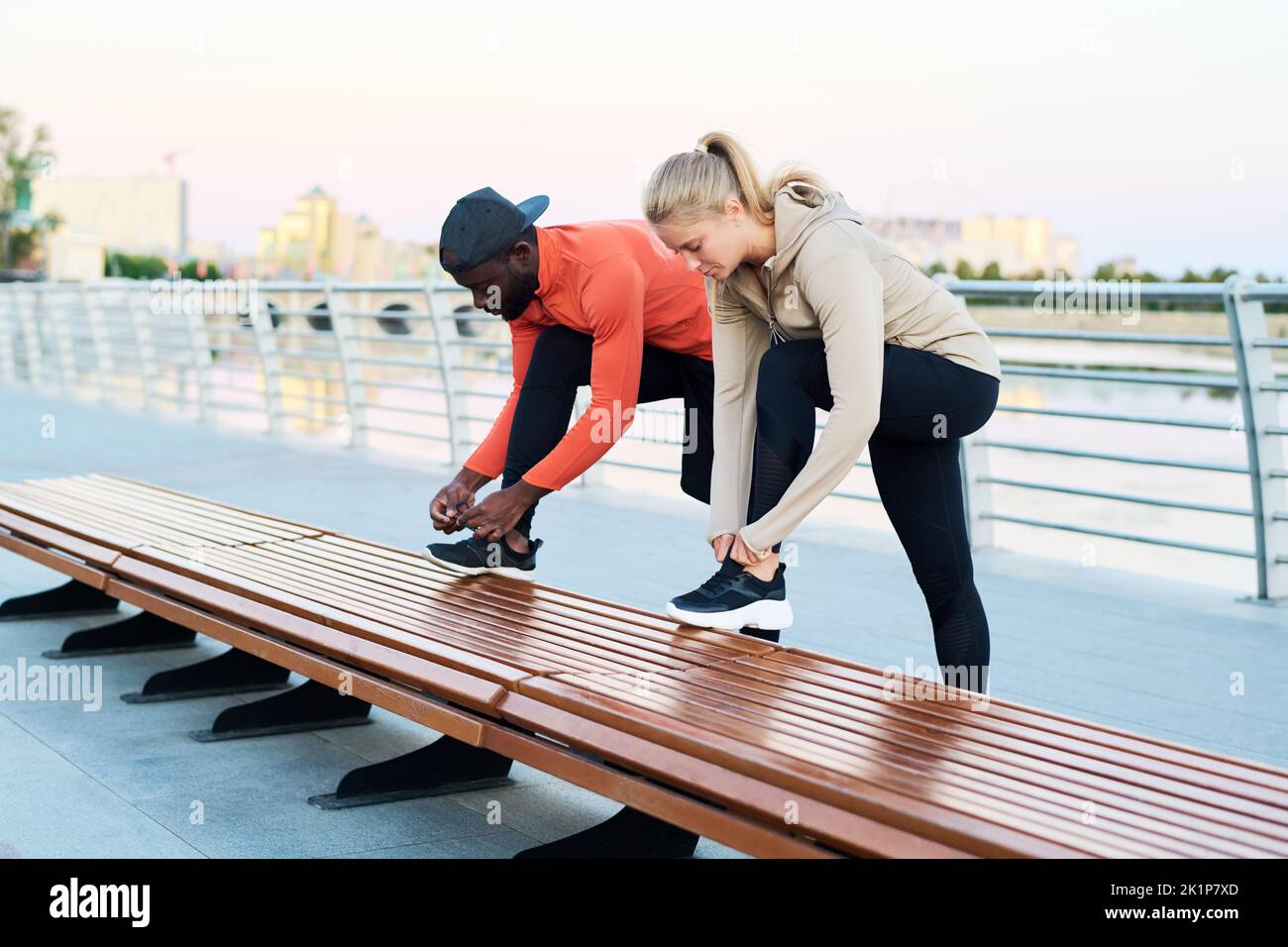 Giovane uomo e donna interculturale in abbigliamento sportivo che legano le scarpe da ginnastica mentre si prepara per l'allenamento all'aperto Foto Stock