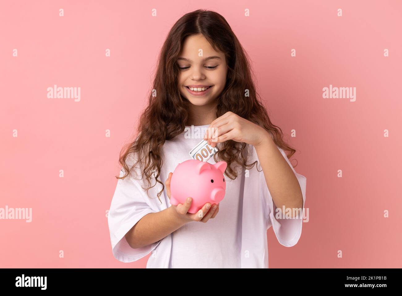 Ritratto di felicissima ragazza sorridente indossando una T-shirt bianca in piedi e mettendo la banconota da dollaro in una banca piggy, risparmiando denaro. Studio in interni isolato su sfondo rosa. Foto Stock