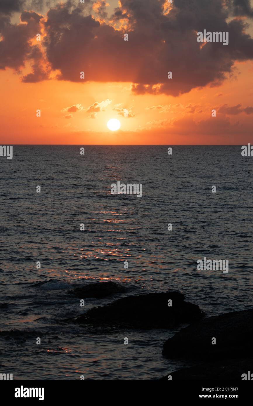 Vista del lontano tramonto dalle scogliere della spiaggia. Colori crepuscolari. Disco solare luminoso in un cielo arancione e nuvoloso. Mare blu. Foto verticale. Idea di fine giornata Foto Stock