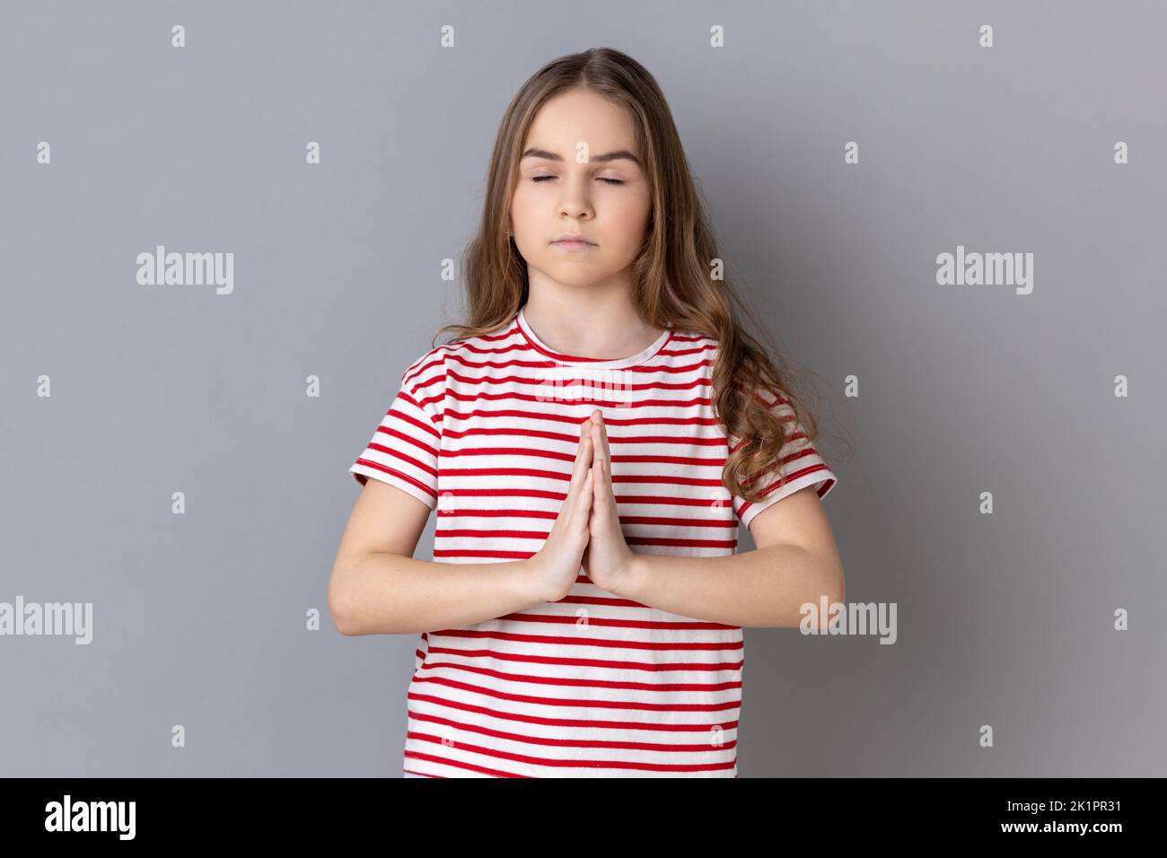 Ritratto della bambina che indossa una T-shirt a righe tenendo le mani gesto namaste, meditare, yoga esercizio tecnica respiro ridurre lo stress. Studio in interni isolato su sfondo grigio. Foto Stock