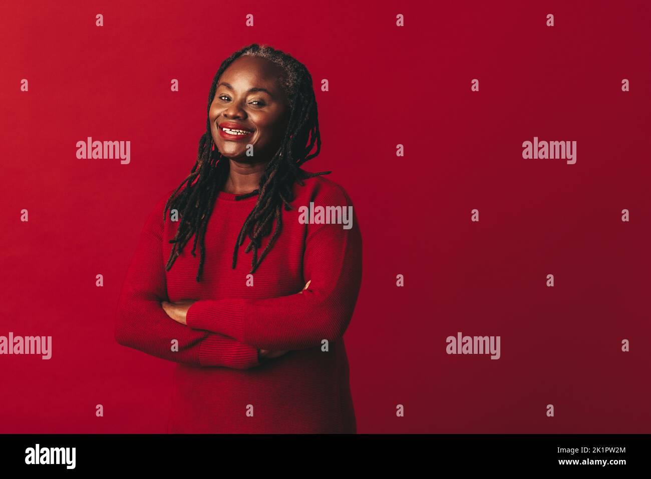 Donna felice con i dreadlocks sorridente alla macchina fotografica mentre si trova su uno sfondo rosso con le braccia incrociate. Donna nera matura che abbraccia il suo natu Foto Stock