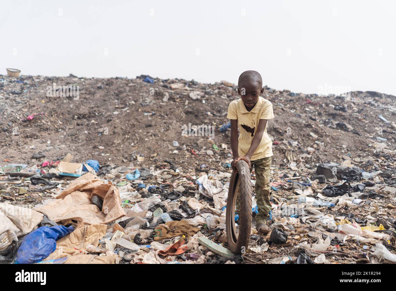 Un ragazzo africano sottopeso che gioca con la sua ruota di bicicletta in mezzo a sostanze plastiche e tossiche in una discarica; rischio di arresto della crescita e trasmissione di malattie in d Foto Stock
