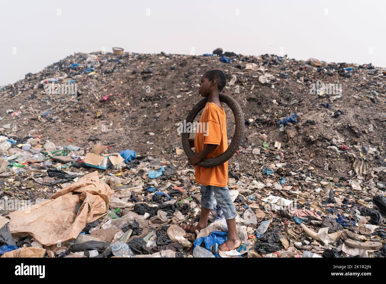 Piccolo ragazzo africano in piedi in una discarica admid mucchi di rifiuti e di sporcizia con una vecchia ruota di bicicletta riciclata sulla sua spalla; concetto di infanzia in poo Foto Stock