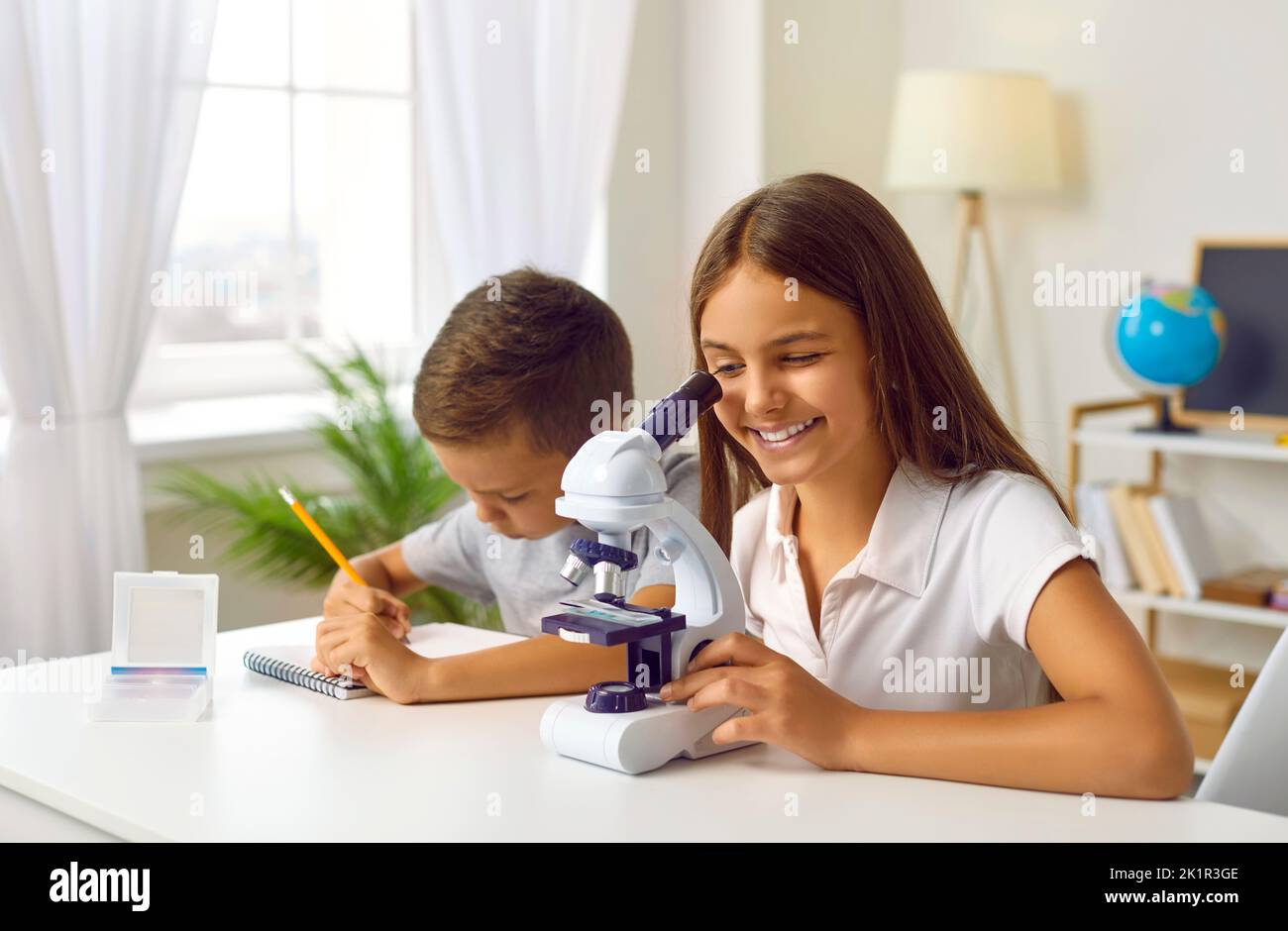 La studentessa sta guardando al microscopio a casa con il fratello minore, preparandosi per una lezione di scienza Foto Stock